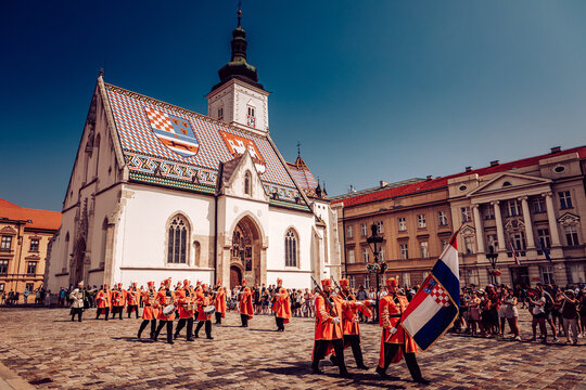 Zagreb, Croatia - 31 August 2019: View of St. Mark's Church with its vibrant tiled roof, as guards in orange uniforms parade with Croatian flag, under a bright blue sky.