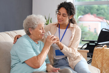 Obraz premium Young African-American female medical worker giving glass of water elderly woman in nursing home