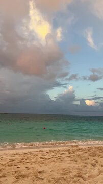 Soft pastel clouds drifting above a calm turquoise ocean along a sandy beach at sunset. 