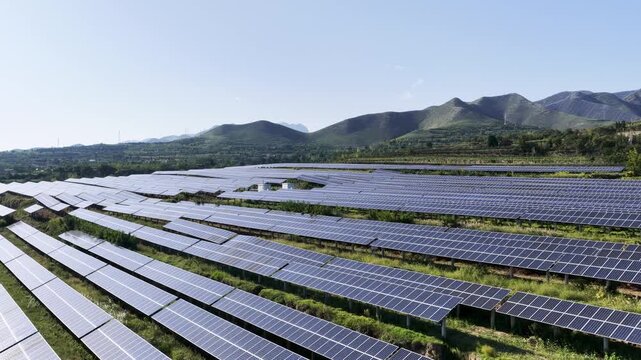 Aerial video of expansive solar photovoltaic arrays stretching across mountain foothills under clear sky, clean power generation, and sustainable rural landscape integration.