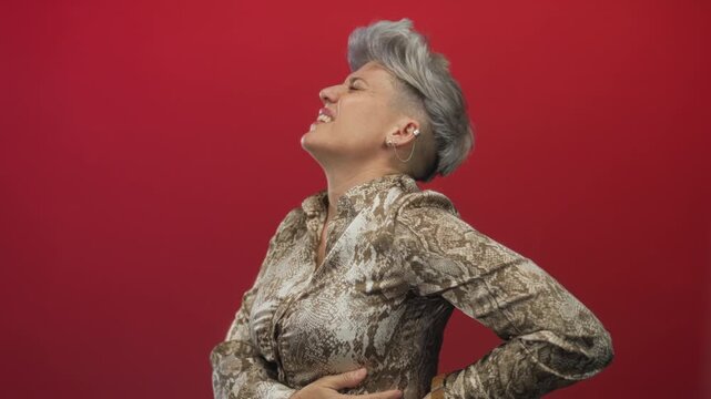 Woman with short silver hair holds lower back in red studio wearing snake print blouse; backache pain.