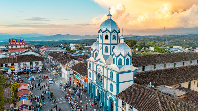 An aerial shot showcases a vibrant blue and white church with a crowd gathered below, set against a dramatic sunset sky.