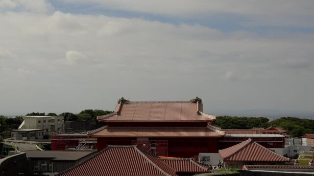 Static high-angle view capturing the distinctive red tiled roofs and traditional Ryukyuan architecture of Shurijo Castle in Naha, Okinawa, Japan, on a cool, cloudy winter day. Tourists walk in the dis