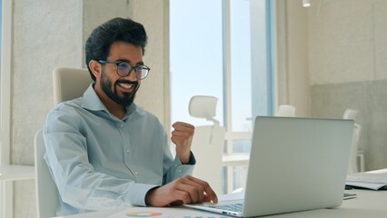 Fototapeta premium Happy Arabian Indian business man celebrating laptop success indoor office excited male guy businessman employer manager triumph winning achievement opportunity winner computer bet deal profit victory