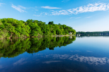 Wide view of a calm Waukesha County lake in June. Vibrant green trees and fluffy white clouds reflect perfectly on the still Wisconsin water.