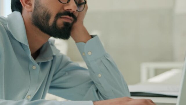 Tired Indian business man sleepy exhausted office worker at workplace bored Arabian entrepreneur leaning hand laptop computer low energy burnout fatigue stress depression job routine exhaustion indoor