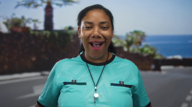 Woman referee blows whistle while gripping her shirt collar on a seaside street with open mouth gesture; surprise.