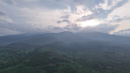 Naklejka premium Serene Mountain Landscape with Layers of Green Hills and Forest under a Cloudy Sky, Sunrays Breaking Through Haze, Panoramic View of Nature's Beauty and Tranquility.