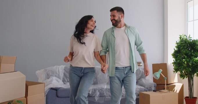 Couple moving to new home apartment, dancing happy dance together in living room interior. Young excited man and woman laugh next to sofa, pile of boxes and lamp, green plant to celebrate relocation
