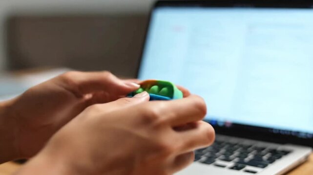 A person using a tactile fidget toy during a professional remote work call, focusing on the hands, soft focus background of a laptop, calm and productive mood