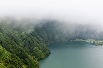 Naklejka premium Volcanic crater lake surrounded by dense fog and green forest in Sete Cidades, San Miguel, Azores. Misty atmosphere with caldera views and serene water.