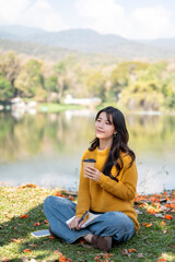 Pretty asian woman holding drinking coffee while sitting crossed legs on grass lawn aside river lake © bongkarn