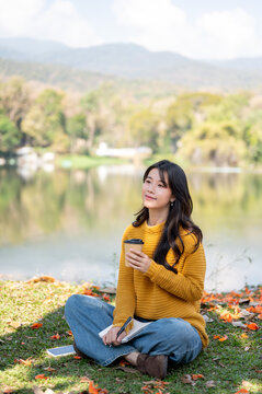 Pretty asian woman holding drinking coffee while sitting crossed legs on grass lawn aside river lake