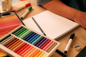 Naklejka premium Angle view of student workspace with blank sketchbook and pencil, art education concept with colorful oil pastels and markers on wooden desk.