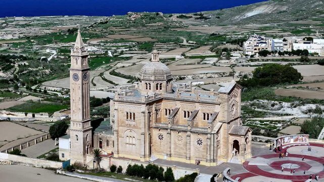 Witness the divine neo-Romanesque Ta' Pinu Basilica in Gozo Malta under a clear summer sky where golden limestone architecture meets scenic green fields in a breathtaking high-angle aerial view.
