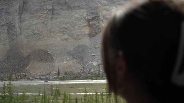 Young woman looks through a bus window and points at rocky mountain scenery during a scenic drive in Canada.