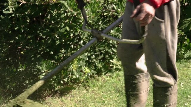 Man wearing a red jacket maintains his garden by cutting weeds and grass with a manual string trimmer.