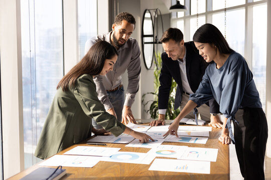 Multiethnic team of analysts working on paper sales reports, analyzing marketing research results, discussing strategy of growth, development, effective business plan, investment