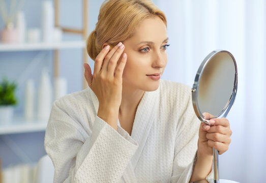 Blonde woman holding round mirror in bathroom to check health of skin and hydration. Skincare beauty routine and home spa treatment of young confident lady in white robe, touching face with fingers