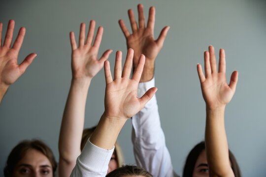 Many hands are raised in unison by an eager group against a simple grey background.