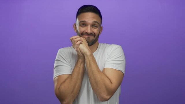 Young man clasping hands with excitement against a vibrant purple background, expressing joy and anticipation in a casual white shirt.
