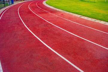 Fototapeta premium Curved red running track with white lane lines beside grass and walkway, ideal for sports facility and race visuals.