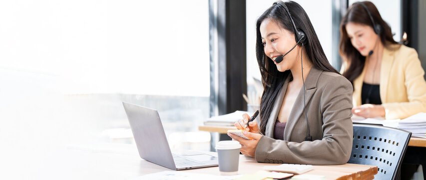 Wide shot of professional Asian customer service team wearing headsets and working on laptops in a bright modern office.