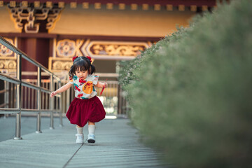 Happy Asian toddler girl in traditional Chinese dress Qipao running and playing with a red envelope (Angpao) at Chinese temple outdoors. Lunar New Year celebration childhood joy and festive atmosphere