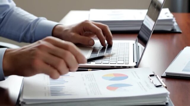 A person's hands typing on a laptop over papers and pens. Close up. Wooden table