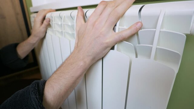 A man's hand touches a white aluminum radiator mounted on a green wall to check the heat. Concept of winter heating season, expensive energy bills, and maintaining home warmth.