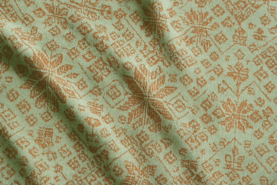 Close-up of soft light green fabric featuring an intricate brown floral and geometric songket pattern. The crumpled texture creates natural folds and shadows, perfect for backgrounds and designs.