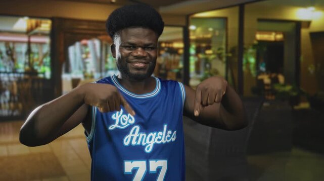 Smiling man points both fingers down at blue los angeles basketball jersey in a brightly lit hotel lobby; confidence.