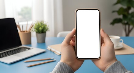 Hands Holding Smartphone With White Screen Above Blue Desk With Laptop And Green Plant