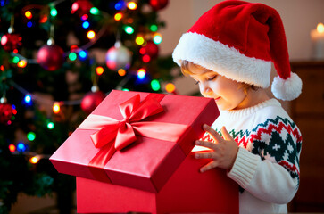 Naklejka premium Caucasian child wearing Santa hat holding large red gift box with ribbon, standing near decorated Christmas tree with colorful lights, looking inside present with curious expression