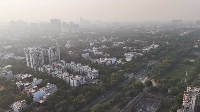 Drone footage of a green roundabout and intersecting roads under a hazy sky, where steady traffic and surrounding buildings reflect rapid urban growth alongside rising air pollution.