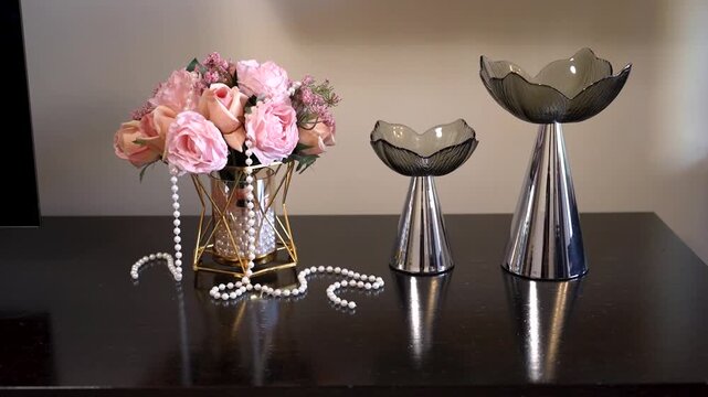 Colorful flowers in a vase sit alongside shiny decorative bowls on a wooden table. Pearls are draped around the vase in a simple home decor setup.