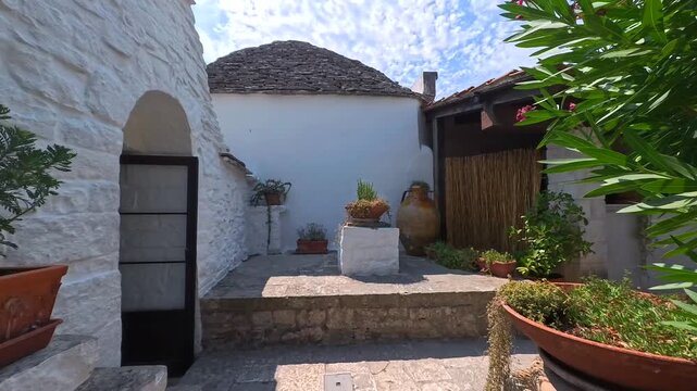 First-person walking through charming courtyard of whitewashed Trullo building with potted plants on sunny day in Alberobello, Puglia, Italy