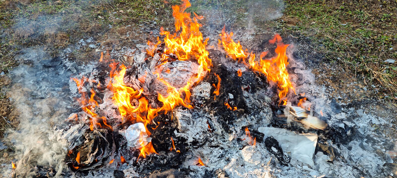 A large outdoor fire burning piles of paper and household waste into ash and smoke on a stone base in a backyard garden