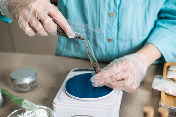Gloved hands pour dark botanical salts from plastic packet into glass test tube on digital scale. Botanical layering, multi-salt DIY, herbal infusion shot, aesthetic bath salts.