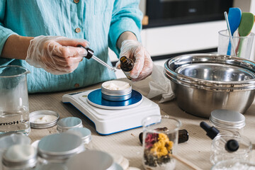 Person uses glass dropper to add oil to white cream in metal jar on digital scale. Lab-style beauty aesthetic, precise cosmetic formulation, diy apothecary lab, scientific skincare diy.