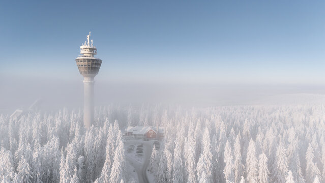 Snow covers the ground and trees around Puijo tower in Kuopio, Finland. Thick fog blankets the area, creating a winter scene. The sky is clear above the tower