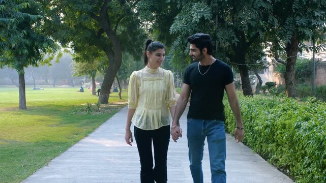 A romantic boyfriend- girlfriend hold hands while walking together on a sidewalk - pedestrians, pavement. A cheerful young romantic couple in the city park - a leisure time, pleasant weather, relation