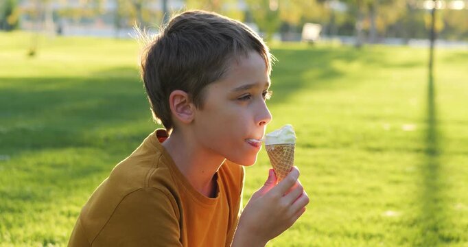 Child boy eating ice cream smiles in summer park at sunset now. Kid eating ice cream holds cone. Warm golden light paints grass. Childhood joy feels simple. Child eating ice cream enjoys sweet treat.