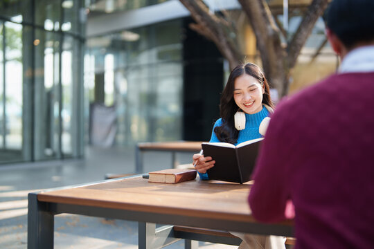 Asian female student smiling and holding an open book, wearing white headphones around her neck, enjoying outdoor learning while sitting at a table on a university campus