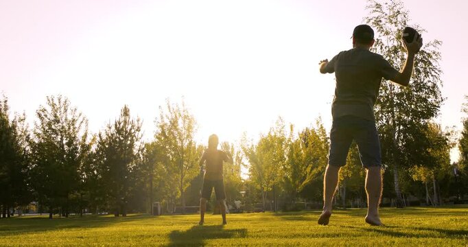 Active father and son play rugby in sunny park. Sporty father throws ball to son at sunset. Happy dad teaches child American football game. Outdoor family fitness and sport concept. Father and child.