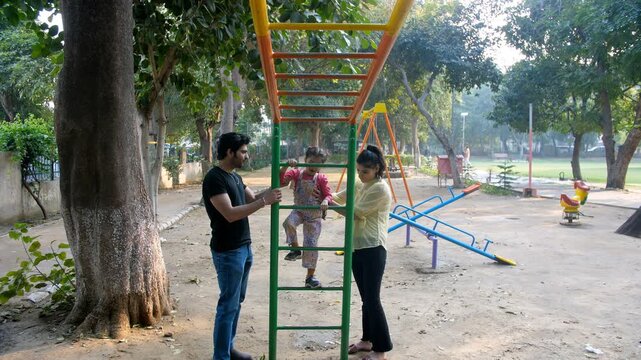 A small little Indian girl having fun climbing on monkey bars - an outdoor exercise, an open playground. An Indian couple at a children's park with their little daughter - rides and swings, leisure