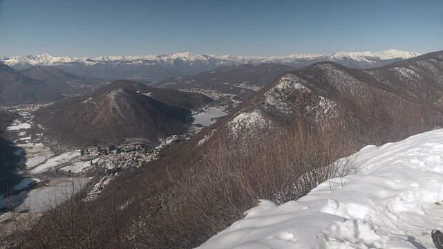 Hiker standing on mountain summit overlooking frozen lakes in winter with Alps in the background. Valganna, province of Varese, Italy. Landscape from Poncione peak