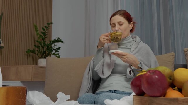A woman sits on a couch with herbal tea in a cup. She holds napkins and tries to recover from her cold while battling symptoms like chills and fever.