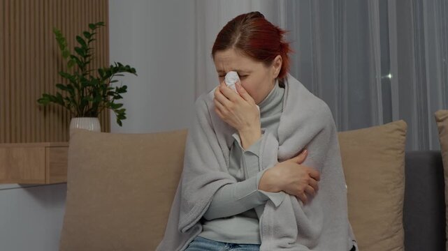 A woman sits on the couch wrapped in a blanket. She sneezes and coughs as she sips herbal tea to feel better while struggling with cold symptoms.