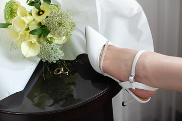 Bride resting her legs in elegant white heels near a window, with a wedding bouquet on a black table. Soft natural light, calm morning atmosphere, bridal preparation concept